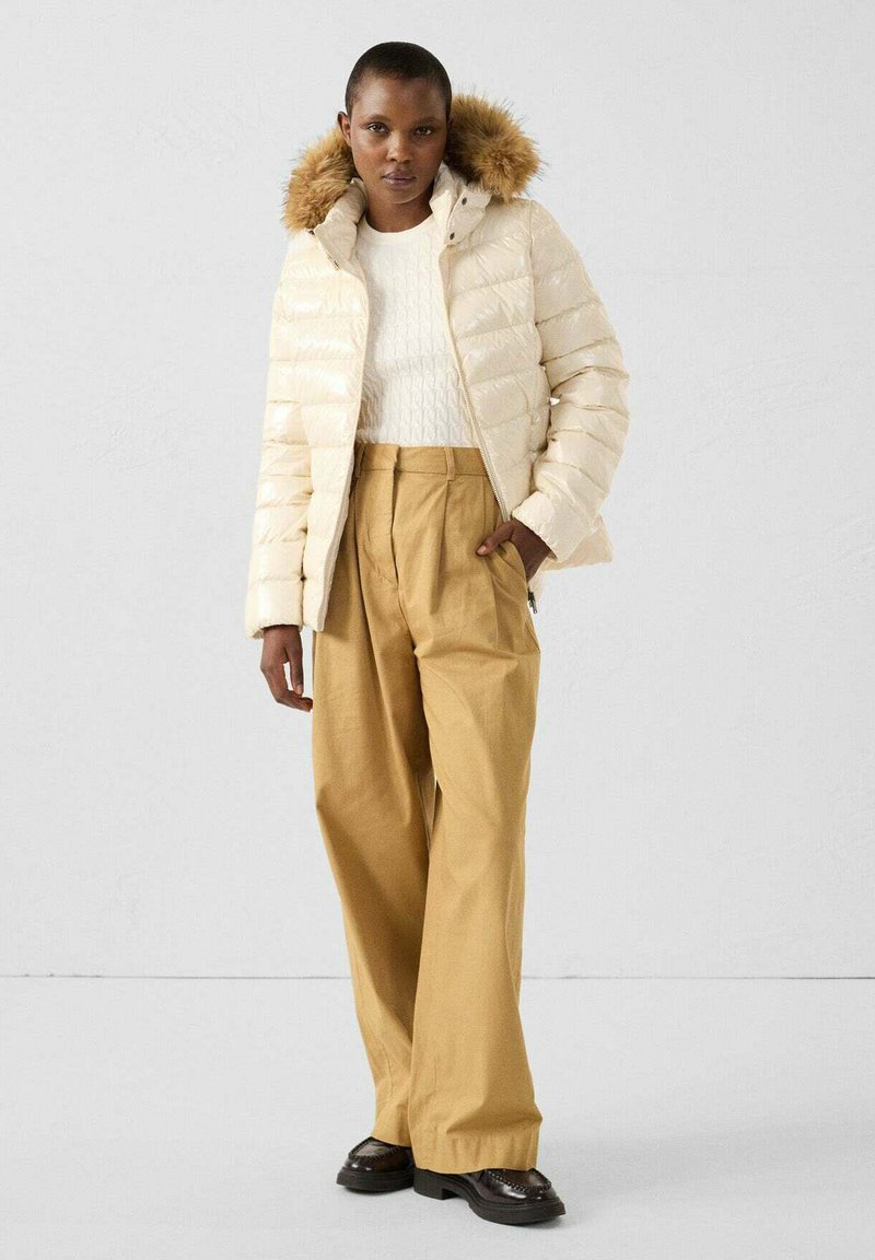 Giubbotto imbottito in beige chiaro con cappuccio in pelliccia sintetica, abbinato a pantaloni beige a vita alta e scarpe nere lucide. Maglione a maglia strutturata sotto.