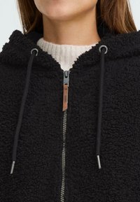 Hoodie en polaire noire avec fermeture éclair à l'avant, présentant une surface texturée, des cordons de serrage et un accent en cuir. Col côtelé visible en dessous.