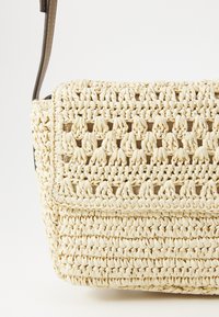 Borsa in paglia intrecciata di colore crema con superficie testurizzata, dotata di chiusura a pattina e una tracolla robusta. Design compatto con una forma strutturata.