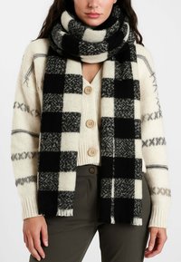 Sciarpa di lana con motivo a quadri bianco e nero, abbinata a un cardigan crema con chiusura a bottoni sul davanti e maniche strutturate.