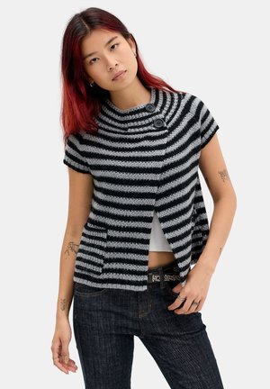 Jeune femme aux cheveux roux portant un cardigan en maille rayé noir et gris à manches courtes et un jean foncé avec des tatouages sur les bras.