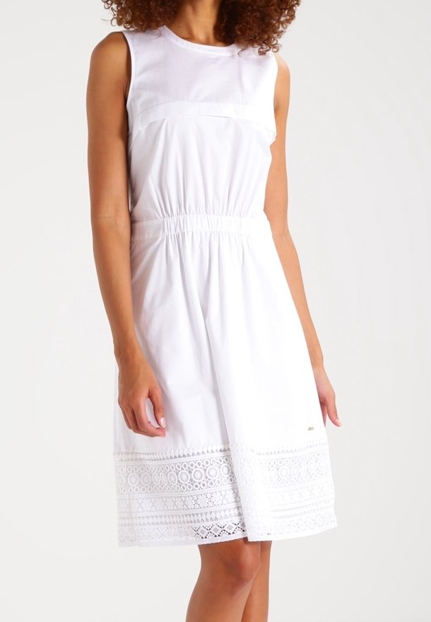 Vestido blanco sin mangas con cintura entallada, que presenta un detalle de dobladillo de encaje y una textura suave. Diseño simple con mínimos acentos de hardware.