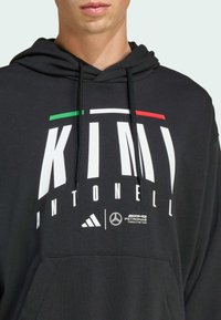 Crna dukserica s prednjim grafičkim isprintom u bijeloj boji i zelenim/crvenim naglascima. Sadrži vezice, kengur džep i logotipe Adidasa i Mercedesa.