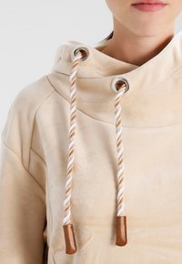Sweat à capuche beige avec une texture douce et veloutée. Présente des cordons torsadés avec des embouts en cuir marron et des œillets en argent.