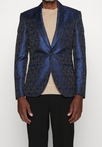 Blazer blu in jacquard con intricati motivi a spirale, chiusura a un bottone, rever a punta e due tasche frontali, indossato sopra un maglione beige.
