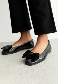 Pies que llevan zapatillas de ballet negras brillantes con grandes lazos de terciopelo negro, combinadas con pantalones cortos negros, apoyados en un suelo moteado.