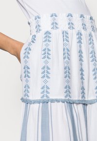 Naisen vyötärö, päällä valkoinen hame, jossa vaaleansinisiä pystysuoria geometrisia kuvioita, rypytetty vyötärönauha, hapsureunus ja käsi taskussa.