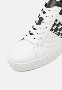 Sneaker bianco con una tomaia in pelle liscia, pannello laterale testurizzato e dettagli neri. Completato con lacci bianchi e una suola in gomma.