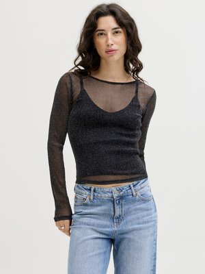 Žena s kučeravými tmavými vlasmi, oblečená v priesvitnom čiernom tričku s dlhým rukávom cez čierny tielkový top a svetlomodré džínsy, stojaca pred jednofarebným pozadím.