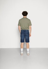 Pessoa com cabelo crespo vestindo uma camisa de listras verdes, calções de ganga azuis, meias brancas e ténis, de pé virada para uma parede branca lisa.