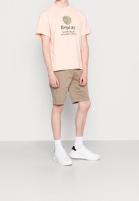 T-shirt rosa a maniche corte con logo stampato, abbinato a pantaloni corti beige e sneakers bianche. Tessuto morbido e vestibilità rilassata.