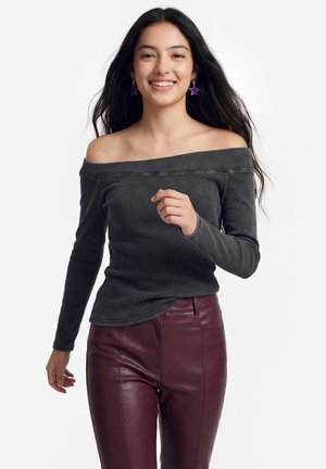 Ung kvinde smilende, iført off-shoulder mørkegrå top, burgunderfarvede læderbukser og lilla stjerneformede øreringe, går fremad.