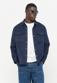 Marine gestreiftes Hemd mit zwei Brusttaschen, aus weichem Stoff. Kombiniert mit einem weißen T-Shirt und blauen Jeans.