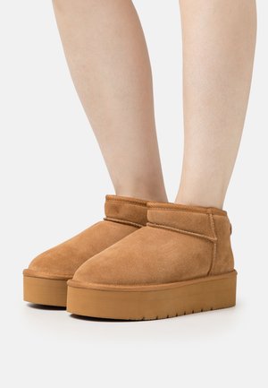 Bottines courtes en daim brun clair avec semelles plateformes épaisses portées sur des jambes nues sur un fond uni.