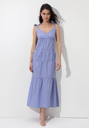 Donna con capelli scuri che indossa un abito lungo senza maniche, lilla chiaro a strati, con laccetti sulle spalle, in piedi scalza su uno sfondo bianco.