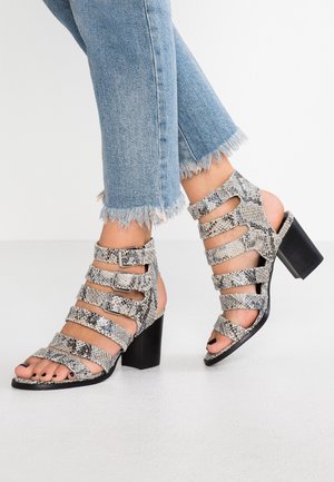 Füße in schlangenmusterbedruckten, riemchenbezogenen High-Heel-Sandalen und hellblauen Jeans mit ausgefranstem Saum auf weißem Boden.