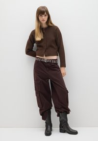 Suéter marrón corto con cierre de botón, combinado con pantalones cargo marrones de corte holgado que tienen bolsillos y un cinturón con tachuelas, además de botas negras hasta el tobillo.