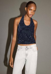 Námornícky modrý crop top so trblietkami a dizajnom za krk, skombinovaný so svetlošedými vysoko pásovými džínsami, ktoré majú gombík a vrecká.