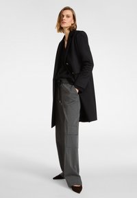 Cappotto nero doppiopetto su una maglietta nera, abbinato a pantaloni cargo grigi con tasche laterali. Scarpa scura a punta completa il look.