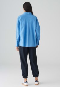 Maglietta a maniche lunghe azzurro chiaro con polsini bottonati, dettaglio con piega nel retro, abbinata a pantaloni jogger blu navy scuri e sneakers bianche con dettagli blu.