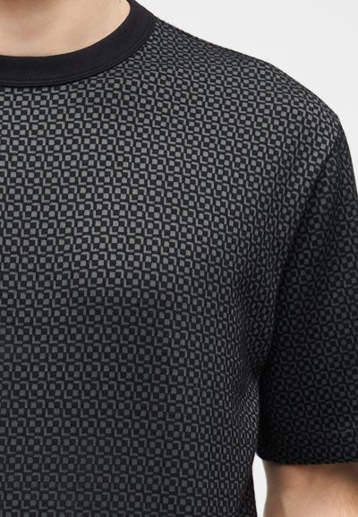 Κοντινό πλάνο ενός κοντομάνικου πουκάμισου με μαύρο και σκούρο γκρι καρό, με στρογγυλή λαιμόκοψη, που φοράει ένα άτομο.