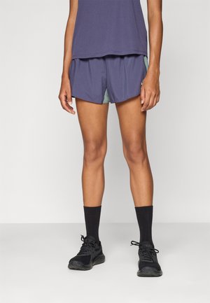 Purple lightweight shorts with a contrasting green inner layer, featuring a relaxed fit and side slits for mobility. Worn with black socks and shoes.