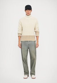 Mand iført sort kasket, cremet ribbet turtleneck sweater, løse falmede grønne bukser og hvide sko, står foran en hvid baggrund.