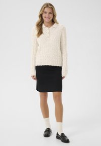 Gestrickter Pullover in Off-White mit einem strukturierten Muster und kragenlosem Ausschnitt, kombiniert mit einem schwarzen Rock und schwarzen Loafers mit cremefarbenen Socken.