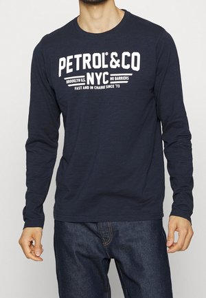 Mężczyzna w granatowej koszuli z długim rękawem z białym napisem "PETROL&CO NYC" i ciemnoniebieskich dżinsach, stojący na tle jasnym.