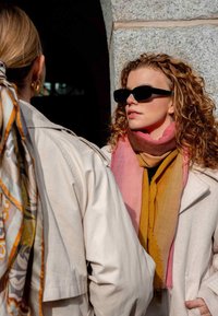 Light pink and yellow gradient scarf worn over a dark top, paired with black sunglasses. The model has curly hair and wears a cream coat.