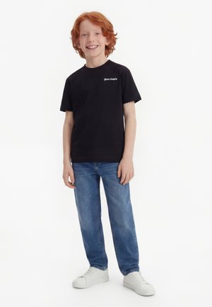 Smiling red-haired boy standing, wearing black “Palm Angels” T-shirt, blue jeans, and white sneakers against a white background.