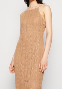 Nainen, joka pukeutuu hihattomaan, beigeen virkatusta neuleesta tehtyyn mekkoon, jossa on korkea kaula-aukko, seisoo yksinkertaista vaaleaa taustaa vasten.