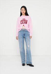 Pulôver rosa de malha com o texto "O CLUBE FRANCÊS" e gráfico de sorvete. Jeans azuis claros com detalhes desgastados. Sapatos pretos.