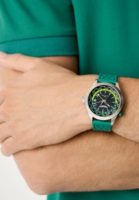 Vyras, dėvintis žalią trumpąsias rankoves marškinėlį, su sukryžiuotomis rankomis, rodo riešo laikrodį su žalia apyranke ir kelių laiko juostų ciferblatu.