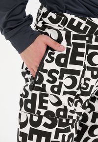 Witte broek met grote zwarte tekstpatronen. Bevat een zijzak met een ritssluiting. Het materiaal lijkt stevig met een lichte glans.