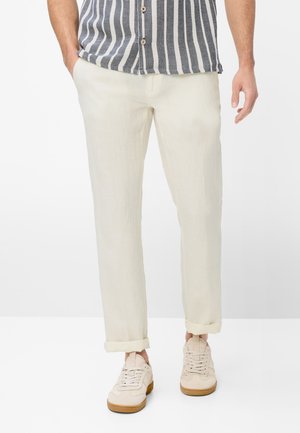 Mand iført beige opknebne bukser, cremefarvede sneakers med gummisåler og en vertikalt stribet skjorte med knapper med hænderne i lommerne.