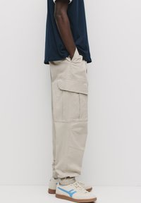 Beige cargo bukser med sidelommer og elastiske manchetter, parret med beige sneakers med blå detaljer og brune såler.
