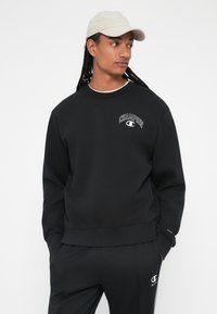 Sweatshirt preto com logótipo "CHAMPION" bordado, decote redondo e mangas compridas. Usado com calças pretas combinando e um boné bege.