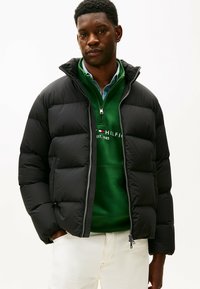 Giacca a vento nera con zip, colletto alto e design trapuntato. Indossata sopra un felpa verde e pantaloni chiari.