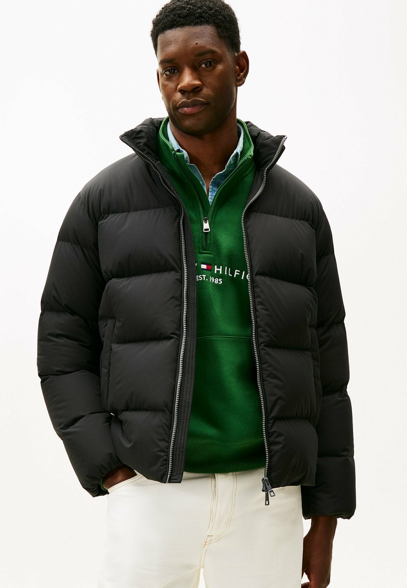 Giacca a vento nera con zip, colletto alto e design trapuntato. Indossata sopra un felpa verde e pantaloni chiari.