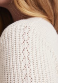 Knit sweater in cream with textured pattern and delicate open stitches along the shoulder. Soft material with a cozy appearance.