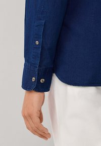 Camicia di jeans a maniche lunghe, colore blu navy scuro, polsini con bottoni e bottoni a contrasto. Tessuto liscio, vestibilità sagomata.