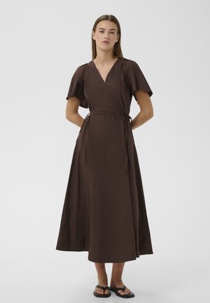 Femme debout, les mains derrière le dos, portant une robe midi marron à col en V, manches courtes, et des sandales plates noires.