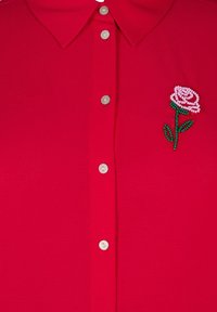 Sarkana blūze ar apkaklīti un pogu slēgšanu priekšpusē. Iezīmējas ar teksturētu audumu un ar pērlītēm izšūtu rozā rozi kreisajā pusē.