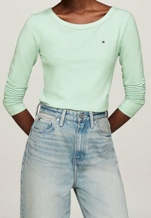 Person iført en lysegrøn ribbet bluse med lange ærmer, stoppet ned i højttaljede lyseblå jeans, med hænderne bag ryggen.