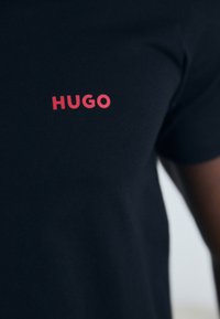 T-shirt navy à manches courtes en coton doux ; arbore un logo "HUGO" rouge vif sur la poitrine gauche. Texture lisse, coupe standard.
