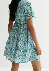 Kukkakuvioinen mekko, jossa on sinivihreä kuvio, lyhyet hihat, solmittava vyötärö ja liehuva helma, joka ulottuu polven alapuolelle.