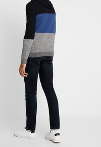 Sweat à capuche en maille texturée avec des blocs de couleur noir, bleu et gris ; doté d'une capuche et de poignets côtelés. Porté avec un jean foncé et des baskets blanches.