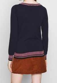 Maglione lavorato a maglia blu navy con collo e polsini a righe rosa e neri, abbinato a una gonna in velluto a coste arancione. Texture liscia, design casual.