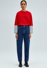 Camisa roja de manga larga con el texto negro "TOUCHÉ", combinada con jeans azules de tiro alto. La modelo lleva botas negras con cordones. Mangas a rayas.